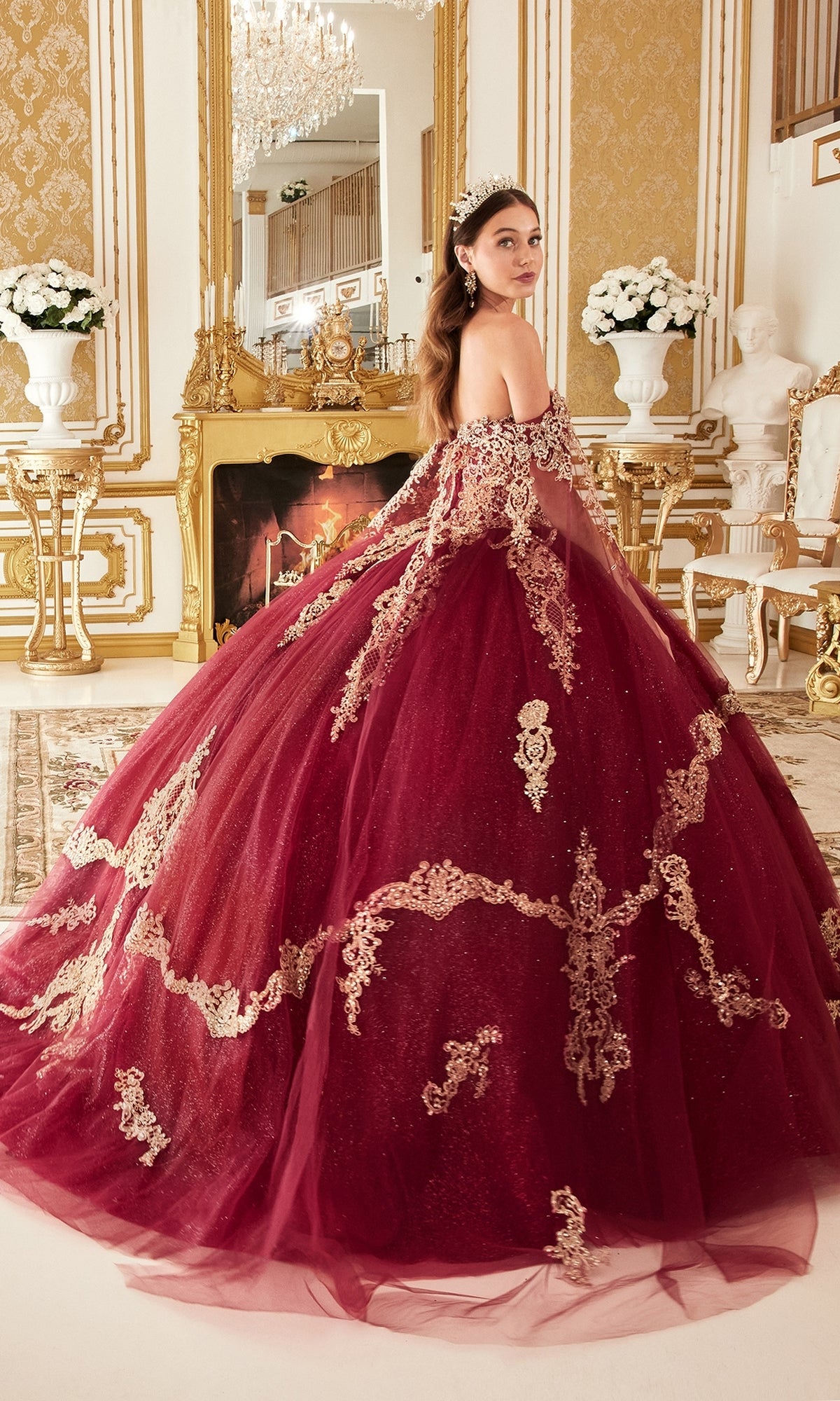 A woman wearing the Ladivine 15711 Quinceañera Dress with gold embroidery stands in an ornate room featuring gold decor, a fireplace, and white flower arrangements.